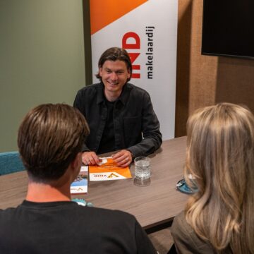 Dick Arts, Makelaar/Taxateur bij VIND Makelaardij: glimlachend in de spreekkamer, met brochure en twee gesprekspartners op de voorgrond.